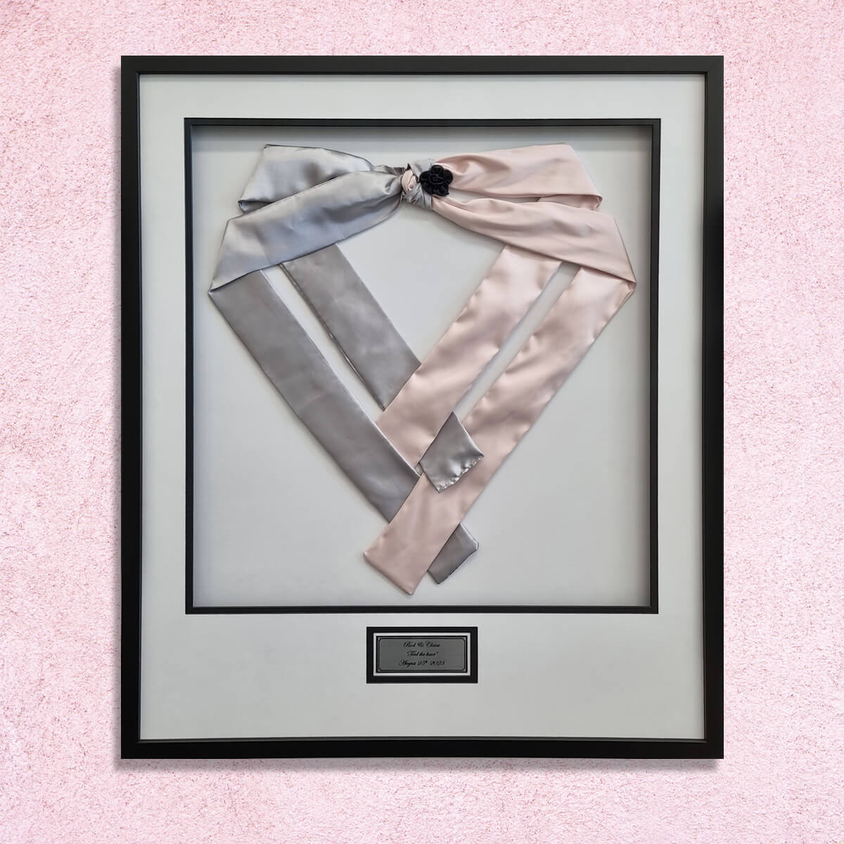 Ceremonial-wedding-sashes-framed-wall Grey and pink satin wedding sashes tied with a black rosette, pinned in a heart shape in a black frame with white double mat and engraved plaque